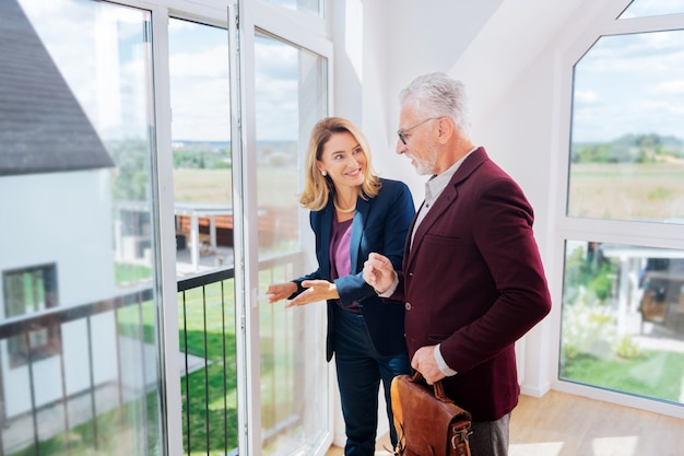 Foto enkele aarzelingen. bebaarde zakenman die elegante jas draagt die aarzelt over het kopen van een nieuw huis dat zich dichtbij makelaar bevindt