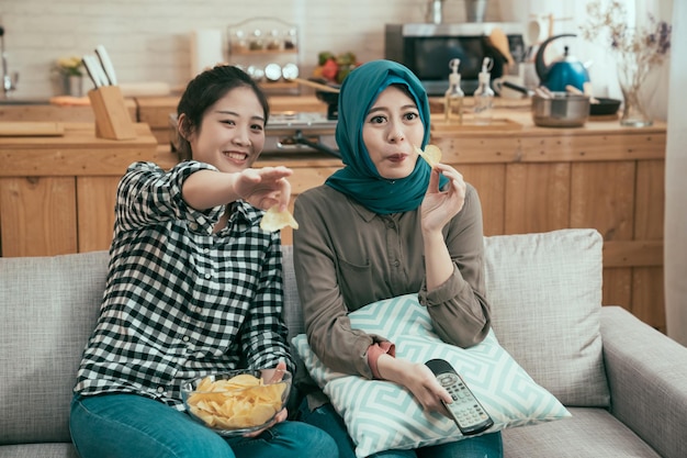엔터테인먼트 및 정크 푸드 개념입니다. 다인종 젊은 여성들은 주말에 아파트에서 함께 자유 시간을 즐깁니다. 한국 소녀는 TV 화면을 가리키고 소파에 앉아 히잡을 쓴 아랍 여성을 보여줍니다.