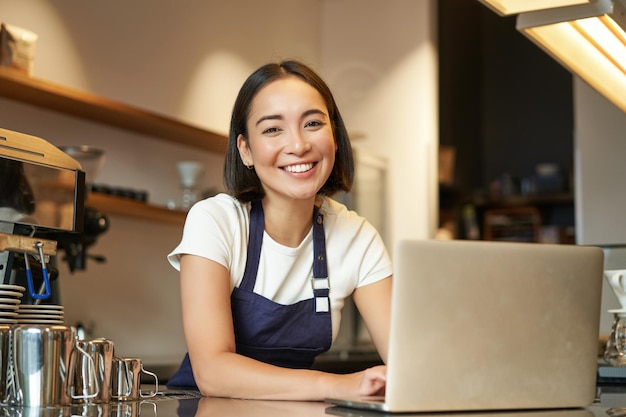 Enthousiast Aziatisch meisje in café-uniforme barista-medewerker met laptop die blij en verrast kijkt naar ca