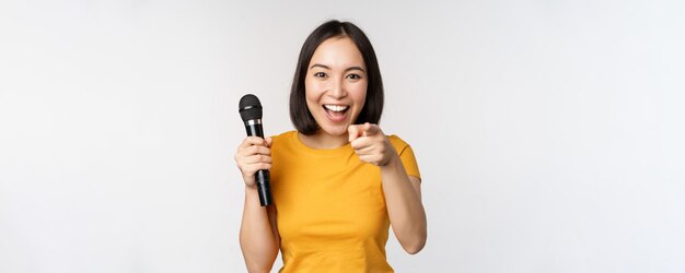 Enthousiast Aziatisch meisje met microfoon die met de vinger naar de camera wijst en suggereert dat je moet zingen terwijl je over een witte achtergrond staat