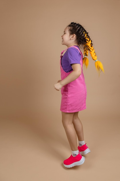 Enthousiast gelukzalig vrouwelijk kind met twee kanekalon-vlechten van gele kleur met plezier springen gekleed in roze jumpsuit paarse tshirt en roze sneakers op beige achtergrond