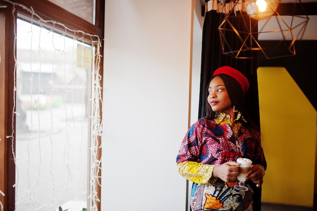 Enthousiaste Afro-Amerikaanse vrouw in trendy gekleurde outfit met rode baret chillen in gezellige café met kop warme latte in handen.