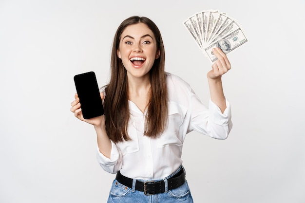 Enthousiaste jonge vrouw die geld wint, met de interface van de smartphone-app en contant geld, microkrediet, prijsconcept, staande op een witte achtergrond.