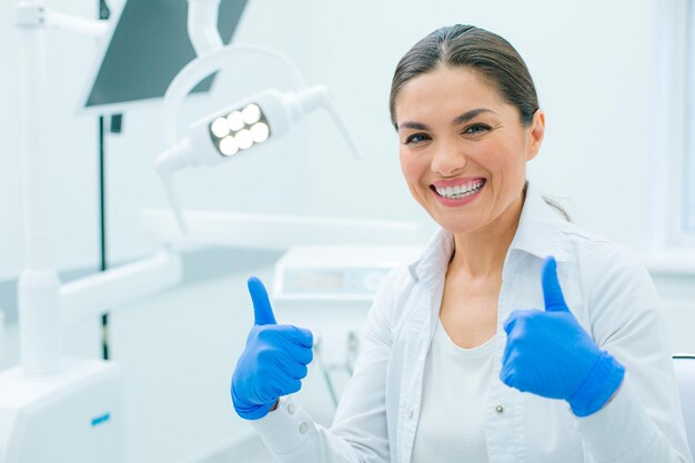 Enthousiaste vrouwelijke arts in een witte jas en rubberen handschoenen die twee duimen opsteken en glimlachen in een kliniek