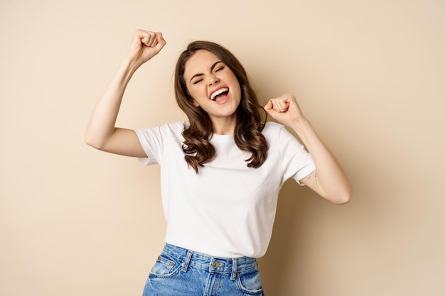 Enthusiastic adn surprised woman winning dancing and celebrating standing in tshirt over beige backg...