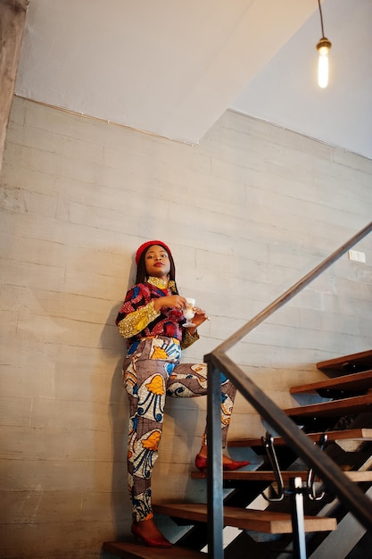 Enthusiastic african american woman in trendy coloured outfit with red beret chilling in cozy cafe standing on stairs with cup of hot drink in hands