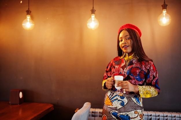 Enthusiastic african american woman in trendy coloured outfit with red beret chilling in cozy cafe with cup of hot latte in hands.