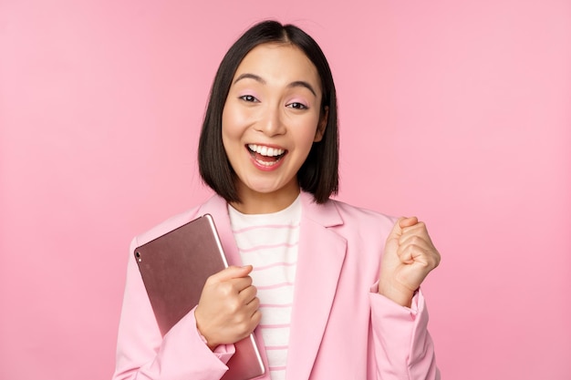 디지털 태블릿을 들고 열광적인 아시아 여성 사업가가 정장을 입고 분홍색 배경 위에 서서 기뻐하며 비명을 지르고 있다