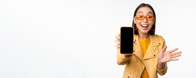 Enthusiastic asian female model showing smartphone app interface online store or website on mobile phone screen standing over white background