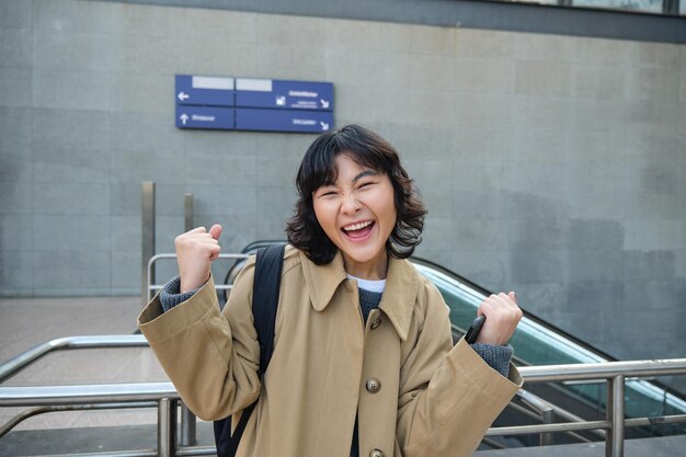 Photo enthusiastic asian girl stands on street makes hooray celebration gesture says yes triumphing winnin