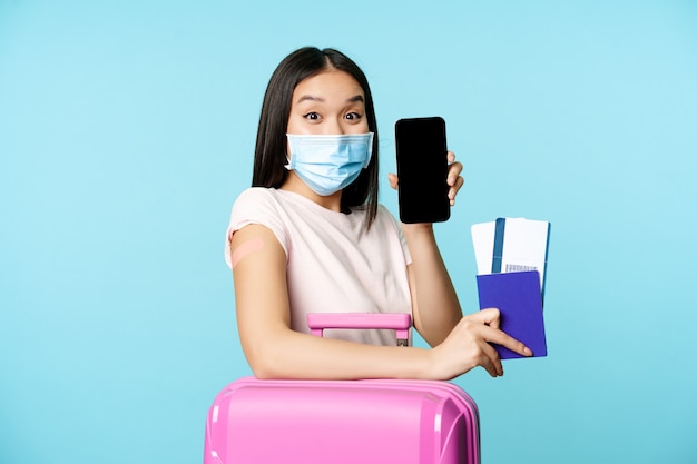 Enthusiastic asian girl vaccinated tourist standing with cute suitcase flight tickets and passport s...