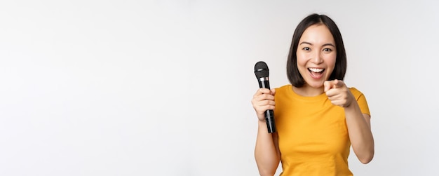 Enthusiastic asian girl with microphone pointing finger at camera suggesting you to sing standing ov...