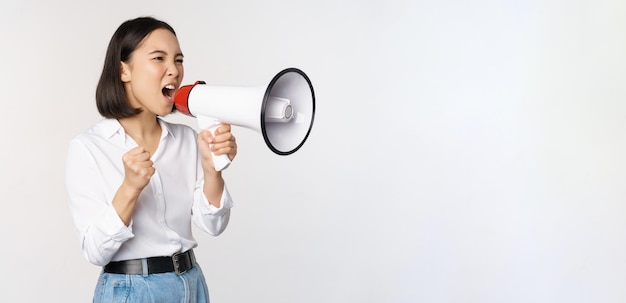 Enthusiastic asian woman girl activist shouting at protest using megaphone looking confident talking in loudspeaker protesting standing over white background