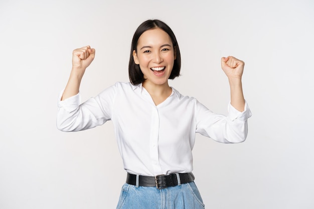 Восторженная азиатская женщина, радуясь, говорит "да", выглядит счастливой и празднует победу, чемпион танцует кулаком, стоя на белом фоне