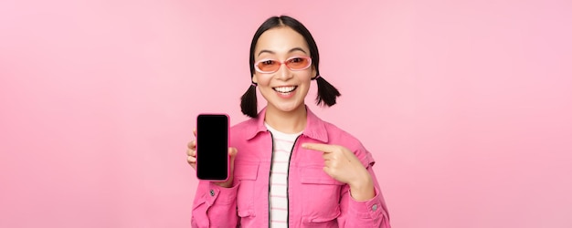 세련된 옷을 입은 열정적인 아시아 여성 선글라스가 분홍색 배경 위에 서 있는 스마트폰 응용 프로그램을 보여주는 휴대 전화 화면에서 손가락을 가리키는