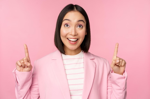 ピンクの背景の上に立っている広告のロゴを表示して指を上に向けて笑顔で熱狂的な企業の労働者アジアのビジネスウーマン