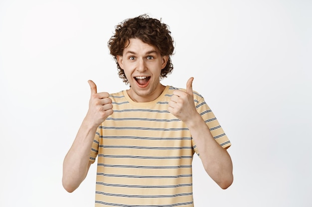 Enthusiastic curly young man showing his support with thumbs up encourage you say yes standing over white background