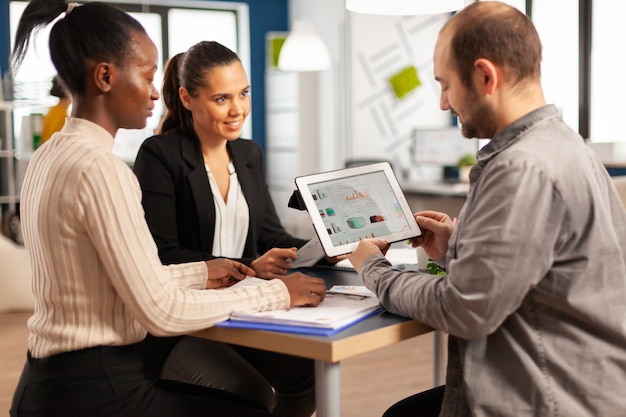 Enthusiastic diverse businesspeople reading annual financial report sitting at table in modern startup business office holding tablet and smiling