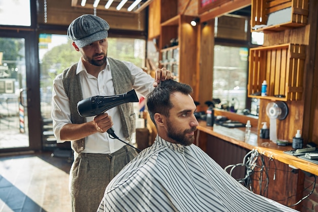 Восторженный парикмахер в модной шляпе сушит волосы феном после мытья.