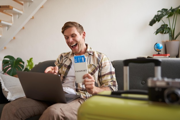 Enthusiastic handsome man tourist holds two flight tickets and passport looks at laptop and screams