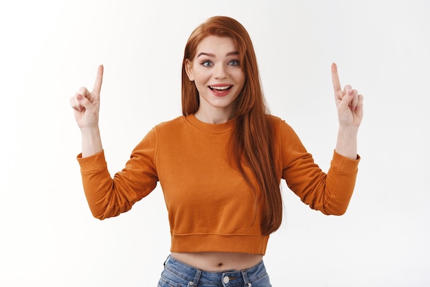 Enthusiastic happy cheerful redhead stylish urban girl wear cropped sweater show belly makeup raise eyebrows excitement joy pointing up index fingers indicate top promo look admiration joy