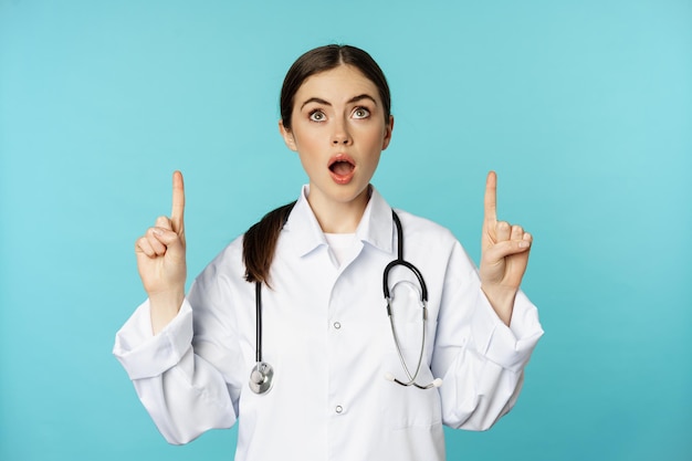 Enthusiastic medical worker, young woman doctor in white coat, stethoscope, showing advertisement, pointing fingers up, standing over torquoise background