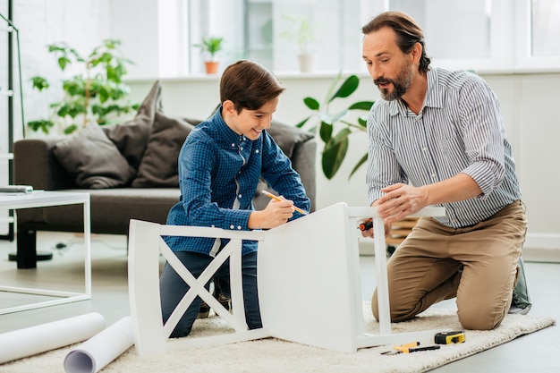 Enthusiastic middle aged man sitting on the floor with his happy teenage son and looking at the white chair while assembling it