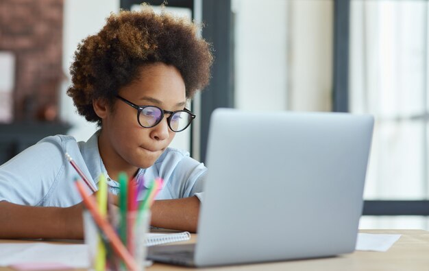 Enthusiastic mixed race teen schoolgirl listening to her teacher while using laptop during online