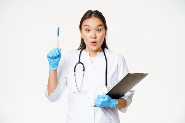 Восторженная медсестра, азиатская женщина-врач поднимает ручку, держит буфер обмена с бумагами пациента, стоит в униформе на белом фоне