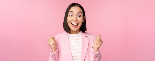 Enthusiastic saleswoman asian corporate woman say yes achieve goal and celebrating triumphing looking with rejoice and smiling standing over pink background