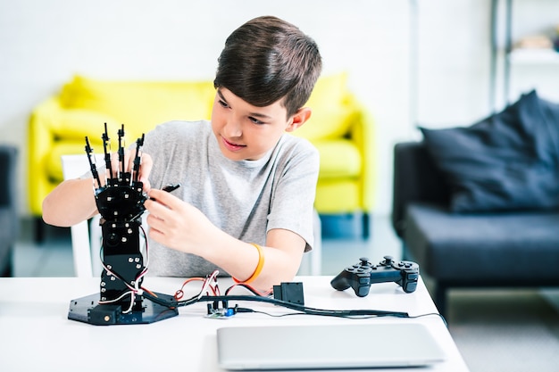 Восторженный умный мальчик сидит за столом, экспериментирует с роботизированной рукой и готовится к инженерным классам