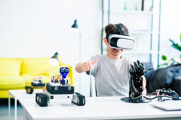 Enthusiastic smart little boy sitting at the table while experimenting with humanoid robotic hand and VR glasses