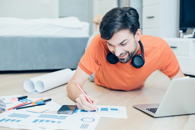 Enthusiastic smiling engineer lying on the floor with a laptop\
in front of him and making notes on colorful graphics