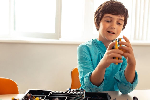 Enthusiastic smiling pupil sitting in the classroom with a box full of equipment in the front and looking for details for his new creation