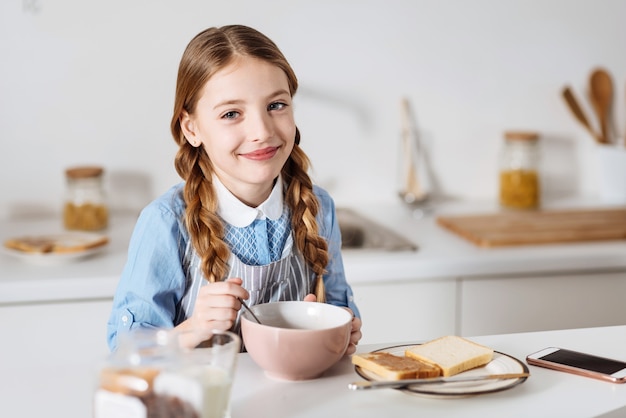 Inizio entusiasta. bambino dolce carino ottimista che si gode il suo pasto mattutino composto da cereali, panini e latte mentre è seduto al tavolo in una cucina