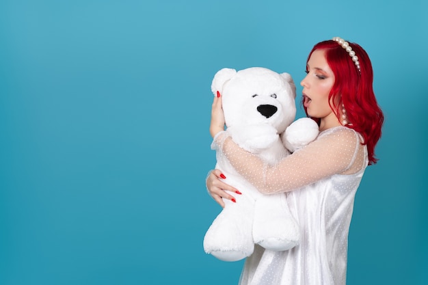 enthusiastic woman in a white dress with red hair and an open mouth embraces a white teddy bear