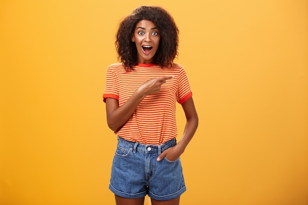 Enthusiastic young African American female with afro hairstyle pointing right with astonished look over orange wall