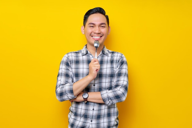 Enthusiastic young asian man wearing a white checkered shirt holding spoon for tasting food isolated on yellow background people lifestyle concept