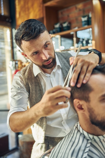 Enthusiastic young barber looking at hair of his client while trimming his hair at the barbershop