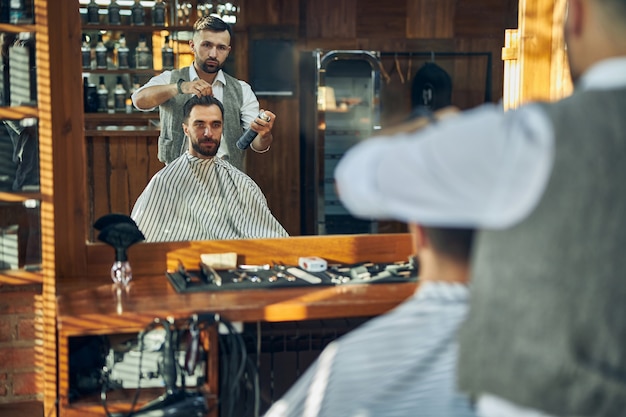 Foto entusiasta giovane barbiere che spruzza i capelli del suo cliente con un fissatore mentre gli dà un nuovo look