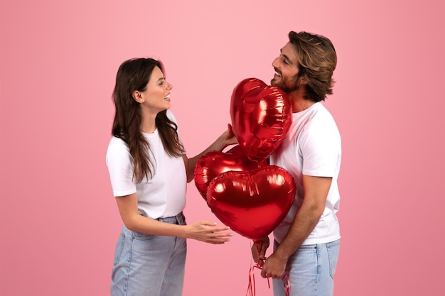 Восторженная молодая пара весело держит и смотрит на блестящие красные воздушные шары в форме сердца вместе