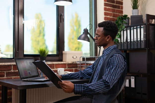 Imprenditore guardando lo schermo del laptop con risultati di vendita che tiene appunti con grafici di affari seduto alla scrivania. impiegato di avvio che confronta gli indicatori chiave di prestazione sul computer portatile.