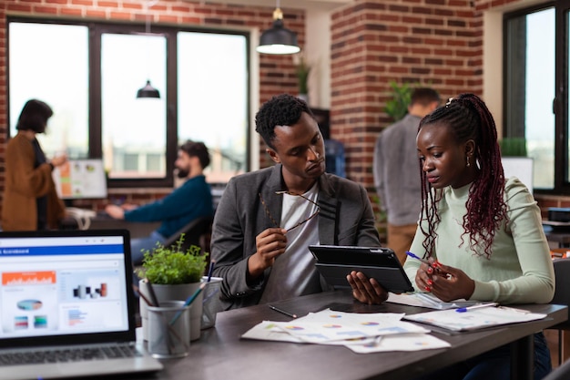 Entrepreneur man holding tablet computer showing company statistics to colleague discussing marketing strategy during business meeting in startup office. Multiethnic team working at financial project