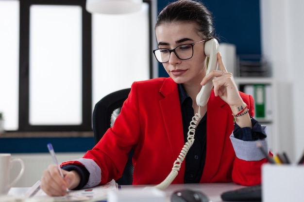 Entrepreneur taking notes in phonecall sitting at desk. Businesswoman with glasses working in startup office. Employee in red jacket writing on paper in telephone conversation.