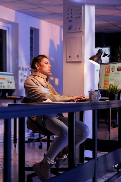 Entrepreneur wearing cervical neck collar in startup office, analyzing financial growth report. Injured corporate employee analyzing statistics typing company strategy late at night in startup office