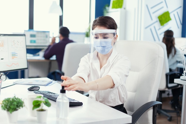 Entrepreneur wearing face mask and face shield using gel for hand hygine during global pandemic with covid-19. Businesswoman in new normal workplace disinfecting while colleagues working in background