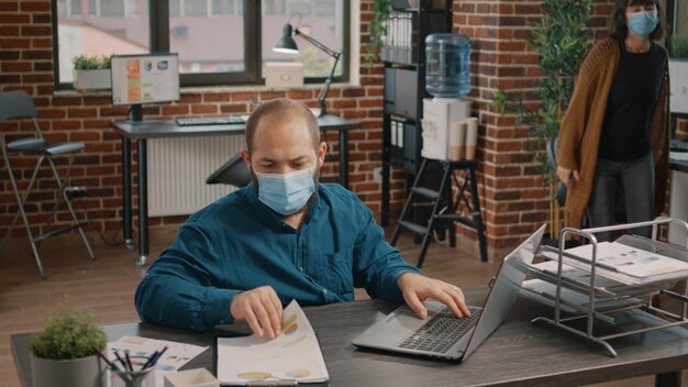 Entrepreneur wearing face mask and working on business project, comparing data rate charts on clipboard and laptop in startup office. Man planning marketing strategy during pandemic