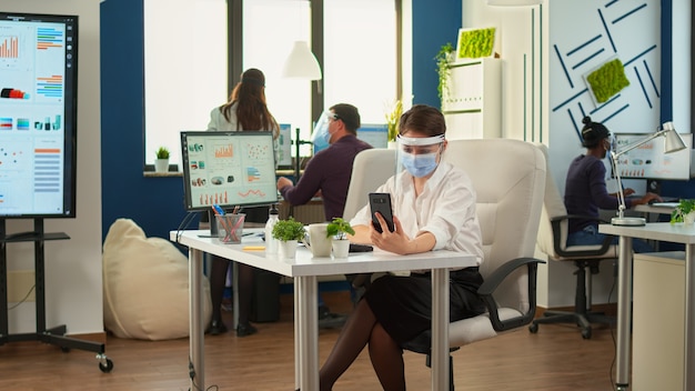 Entrepreneur wearing visor against coronavirus using phone with wireless headphone for videocall talking with remotely team sitting in new normal office. Colleagues working respecting social distance