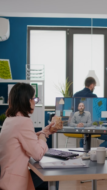 Entrepreneur woman sitting at desk in company office eating\
sandwich during online videocall confere...