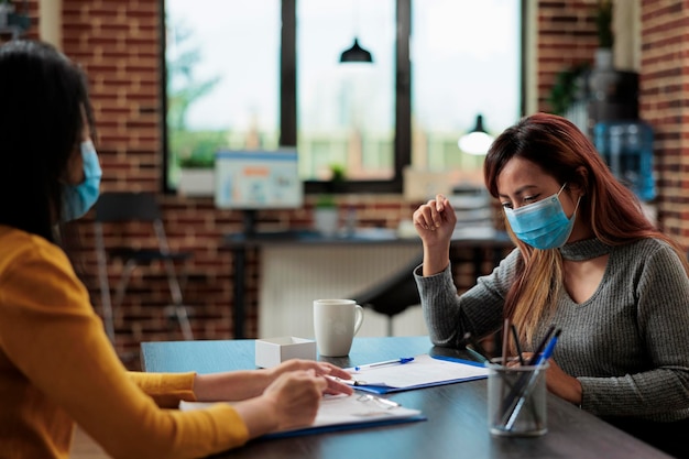 スタートアップオフィスの事業計画で働いているコロナウイルスに対して医療用フェイスマスクを着用している起業家の女性。管理戦略について話し合うマーケティングプロジェクト中に協力する同僚